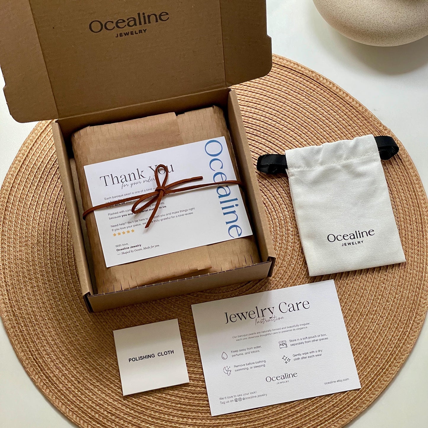 Ocealline jewelry box with jewelry care card, polishing cloth, and jewelry bag on a woven mat.