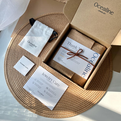 Ocealine jewelry packaging with a cardboard box, jewelry care card, and branded pouch on a woven mat.