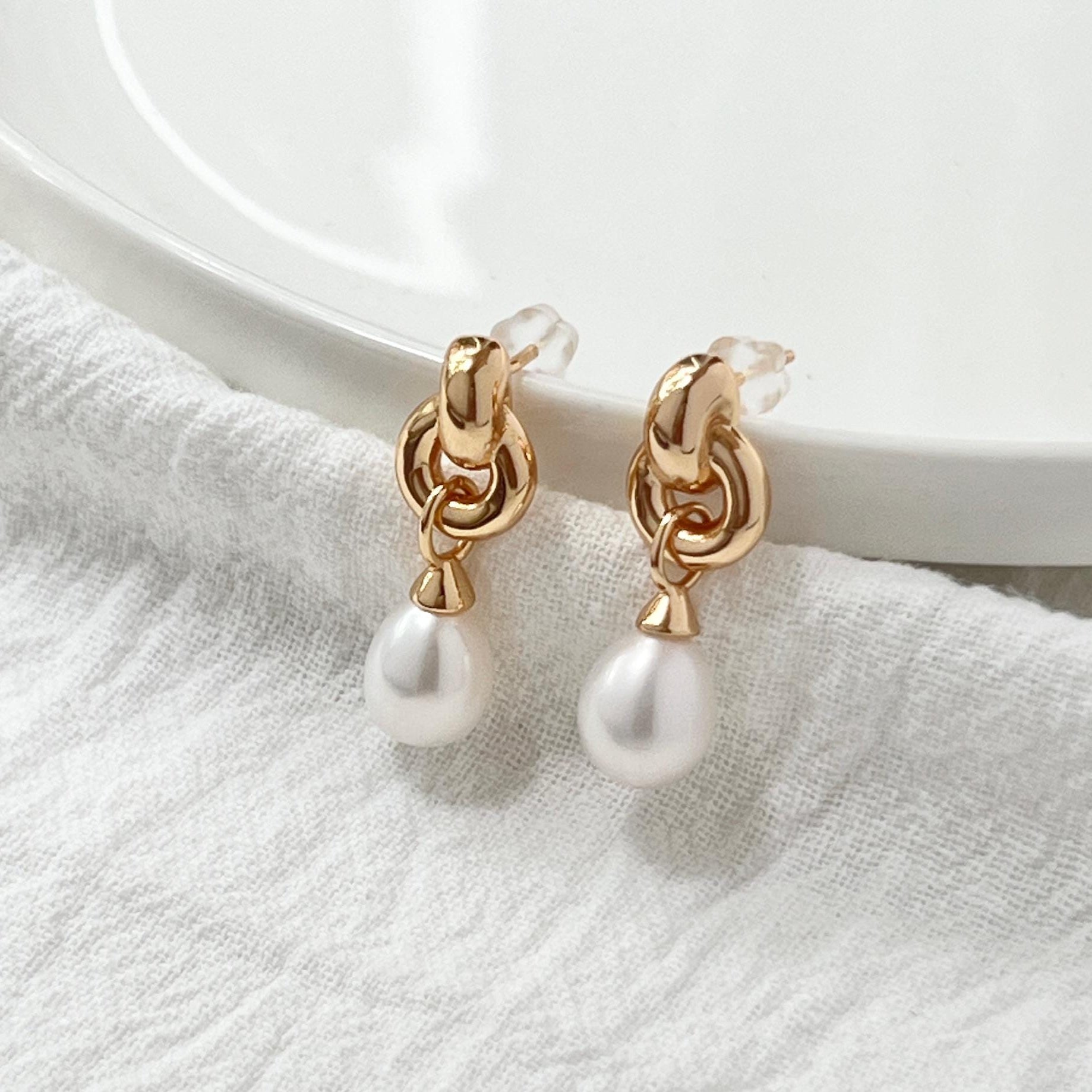 May include: A pair of gold-colored earrings with a dangling pearl. The earrings have a knotted design at the top and a small, round pearl hanging from each. The earrings are displayed on a white, textured surface.
