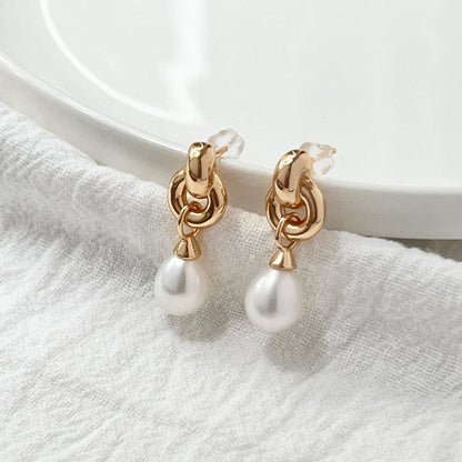 May include: A pair of gold-colored earrings with a dangling pearl. The earrings have a knotted design at the top and a small, round pearl hanging from each. The earrings are displayed on a white, textured surface.