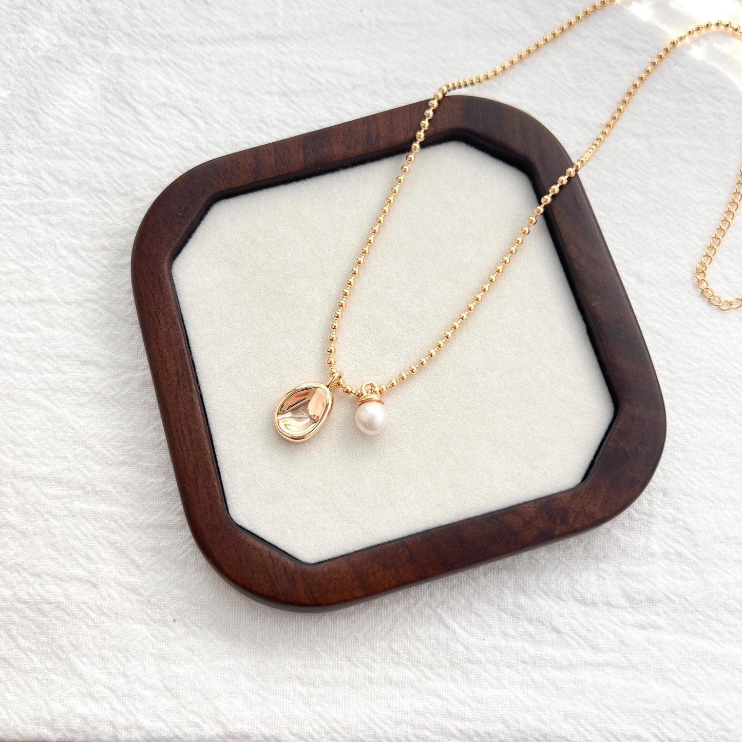 May include: A gold necklace with a pendant and a pearl. The pendant is an oval-shaped, clear stone with a gold border. The necklace has a delicate chain with small, round beads. A small, white pearl hangs next to the pendant. The necklace is displayed on a wooden tray.