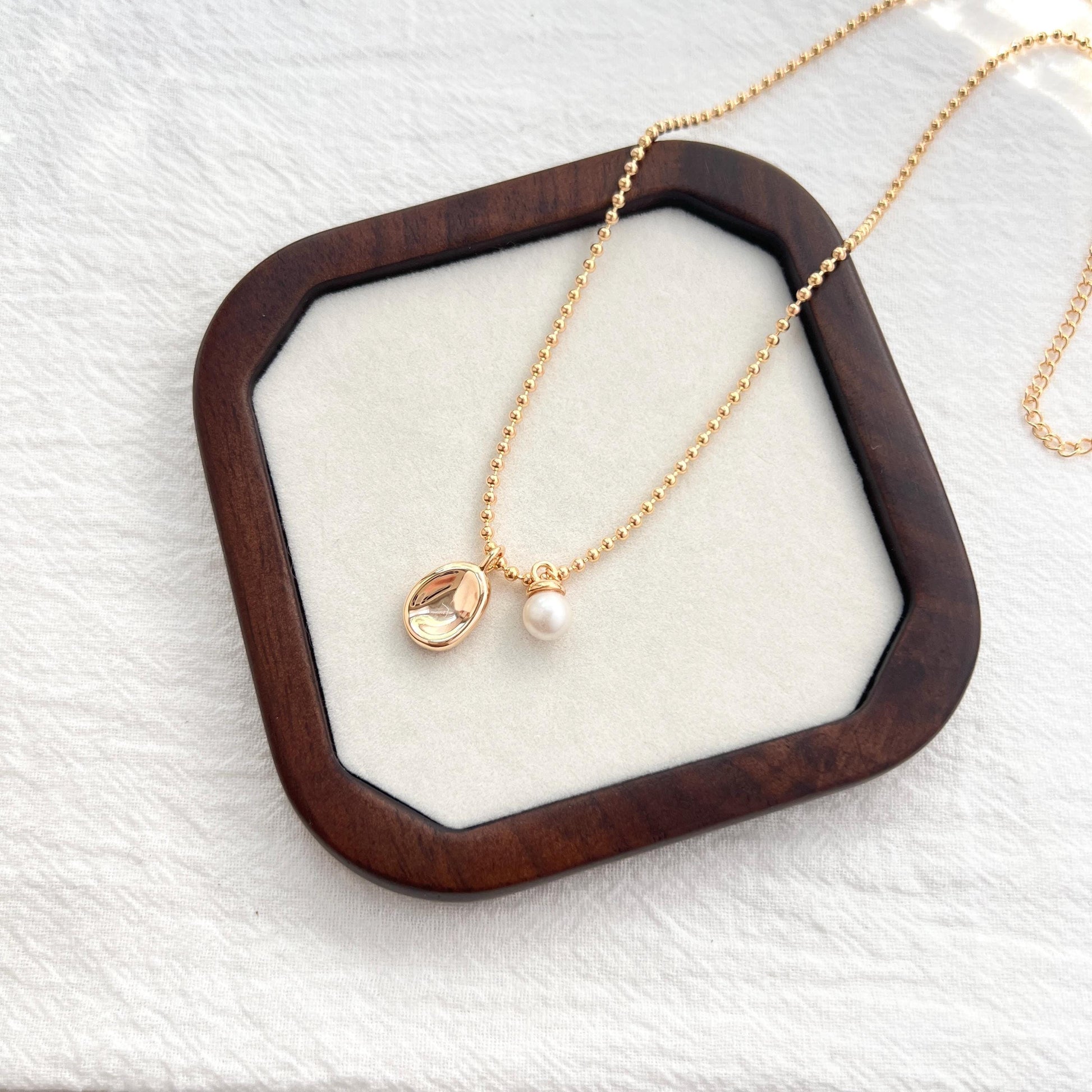 May include: A gold necklace with a pendant and a pearl. The pendant is an oval-shaped, clear stone with a gold border. The necklace has a delicate chain with small, round beads. A small, white pearl hangs next to the pendant. The necklace is displayed on a wooden tray.
