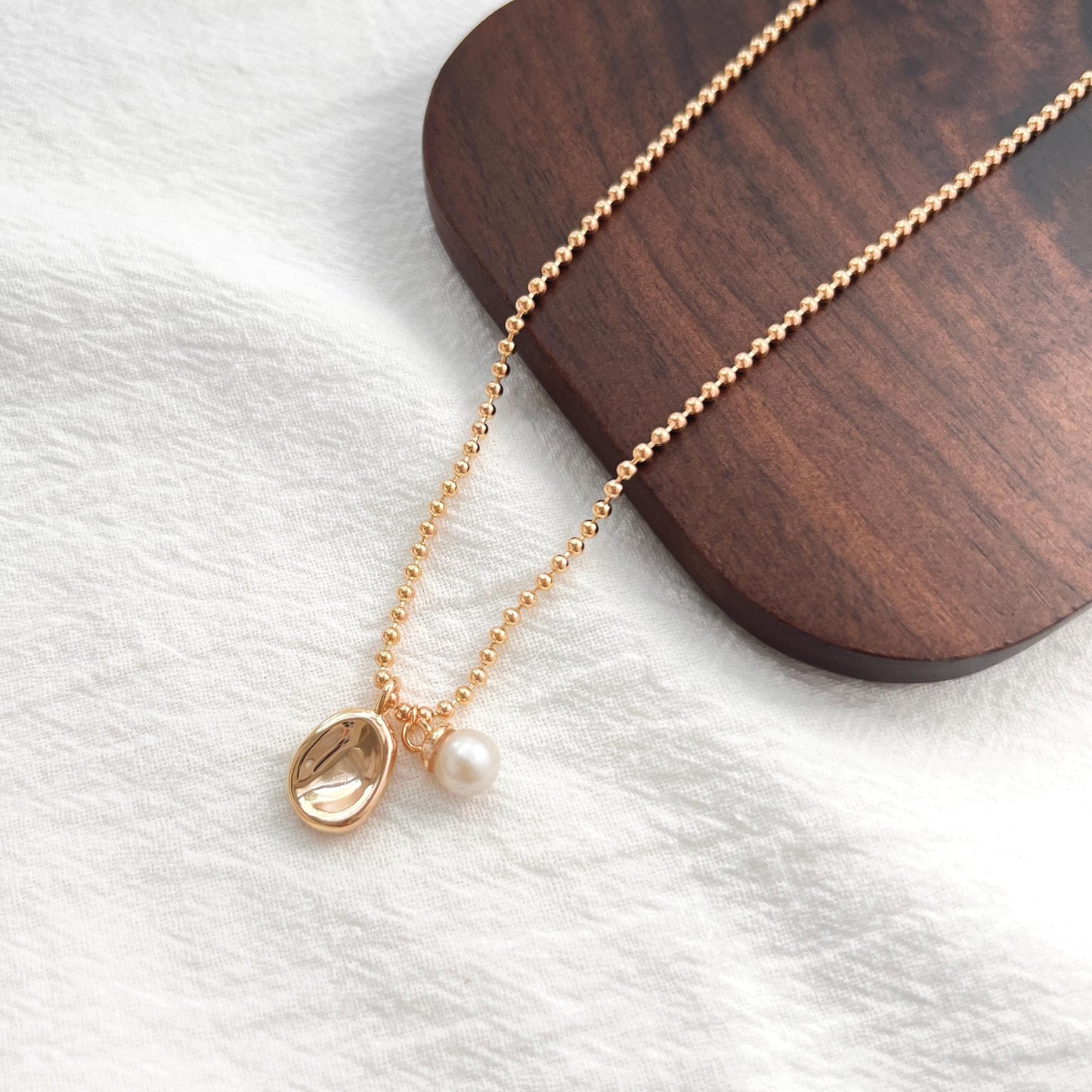 May include: A gold-colored necklace with a pendant and a pearl. The necklace chain is made of small, round beads. The pendant is an oval shape with a textured surface. A small, white pearl hangs below the pendant.