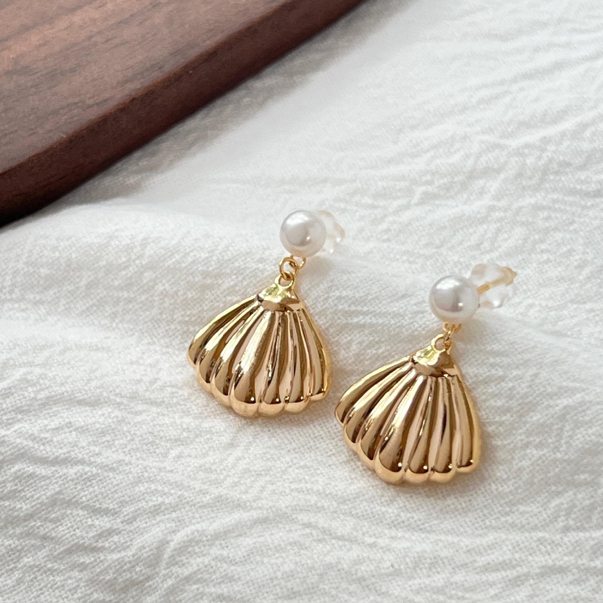 May include: A pair of gold-colored seashell earrings with a pearl accent. The earrings have a detailed, ridged design and a small, round pearl at the top. The earrings are displayed on a white, textured surface.