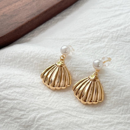 May include: A pair of gold-colored seashell earrings with a pearl accent. The earrings have a detailed, ridged design and a small, round pearl at the top. The earrings are displayed on a white, textured surface.