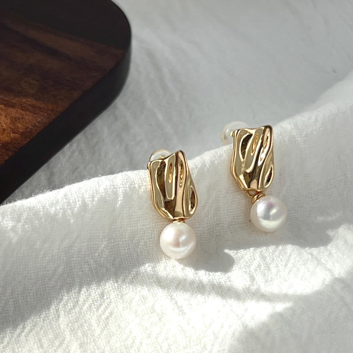 May include: A pair of gold-colored earrings with a textured, rectangular design. Each earring features a small, dangling white pearl. The earrings are displayed on a white, textured surface, with a dark wooden object in the background.