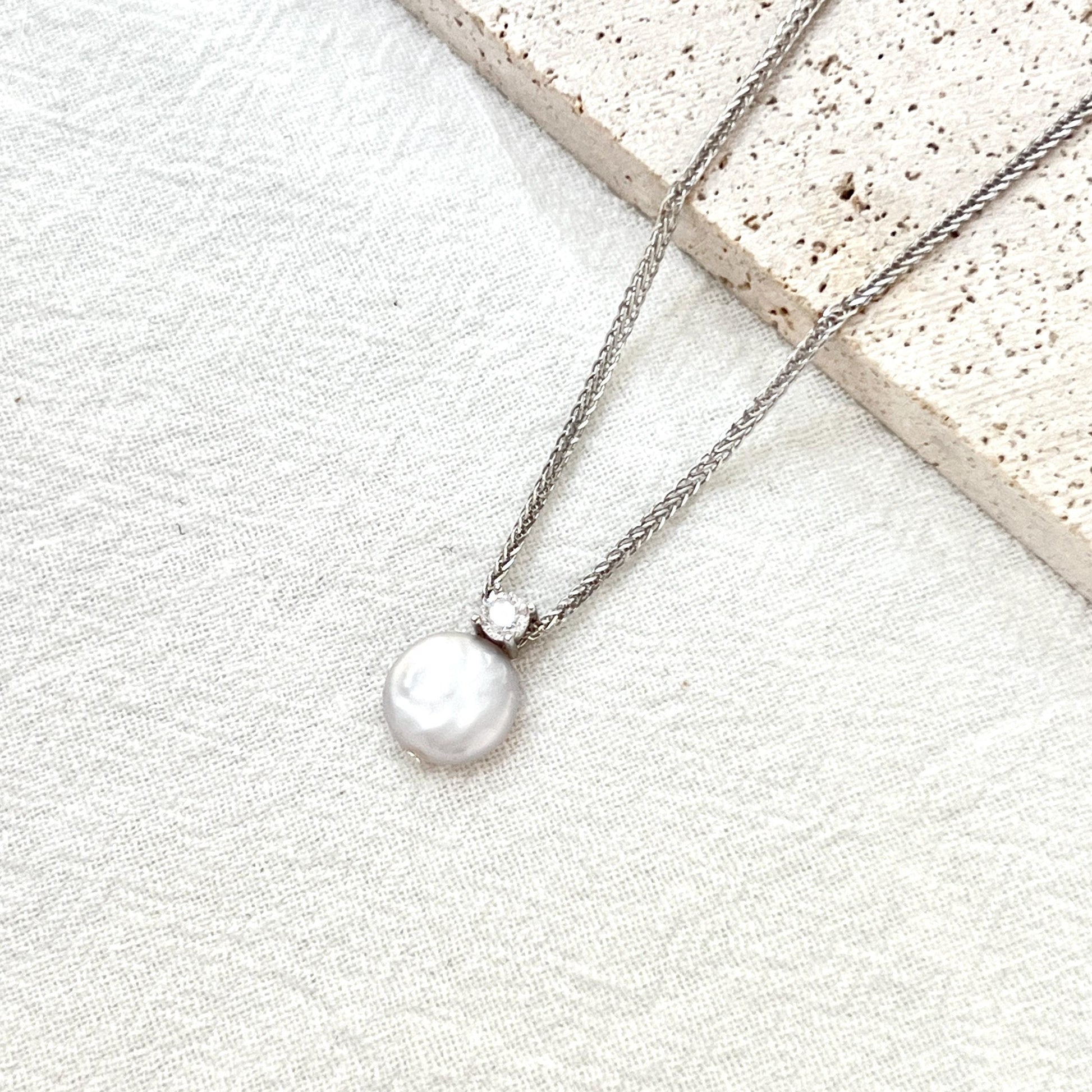 May include: A silver necklace with a round, white pearl pendant. The pearl is connected to the chain by a small, clear gemstone. The necklace is displayed on a white, textured surface, with a light-colored stone in the background.