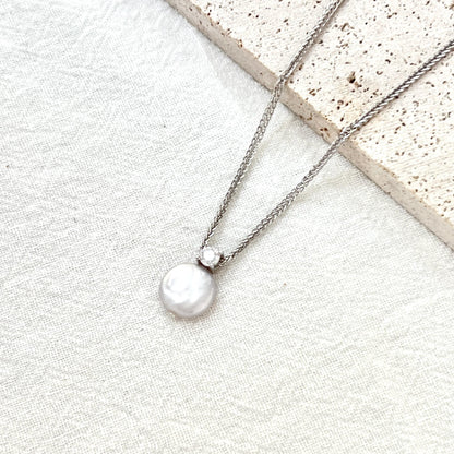 May include: A silver necklace with a round, white pearl pendant. The pearl is connected to the chain by a small, clear gemstone. The necklace is displayed on a white, textured surface, with a light-colored stone in the background.