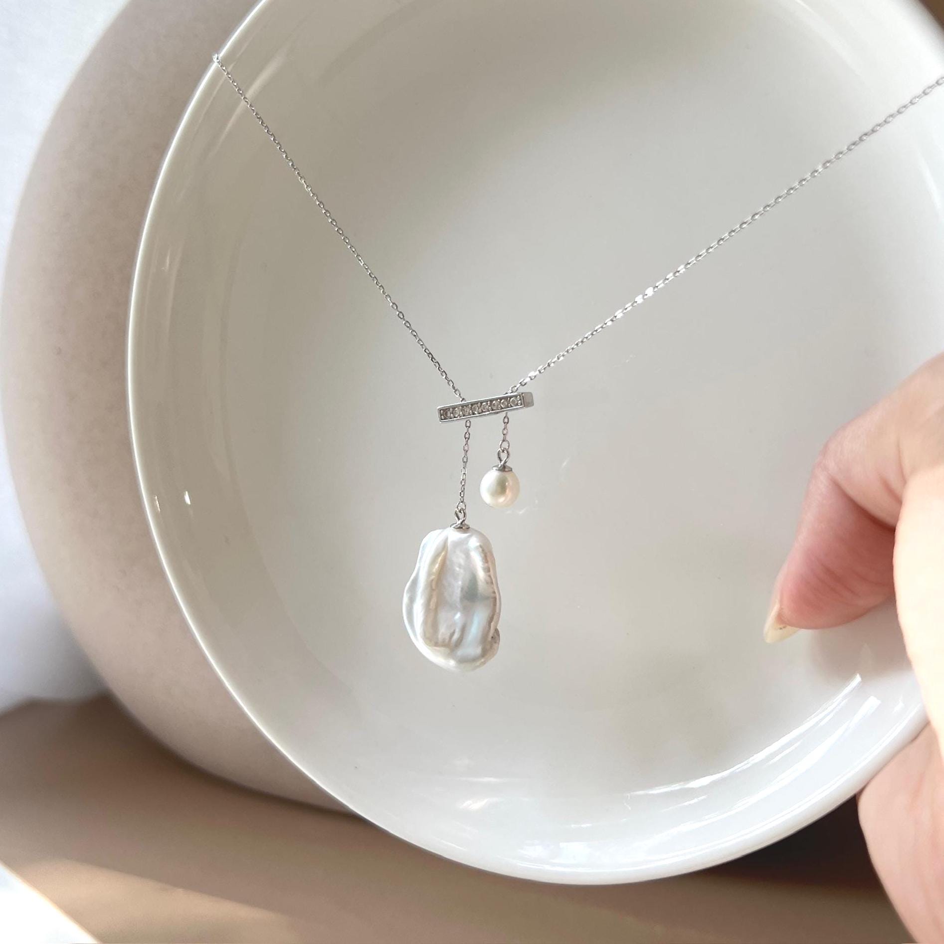 May include: A silver necklace with a bar pendant and two pearl drops. The bar is adorned with small, clear stones. One pearl is a small, round bead, and the other is a large, irregularly shaped white pearl. The necklace is displayed on a white surface.