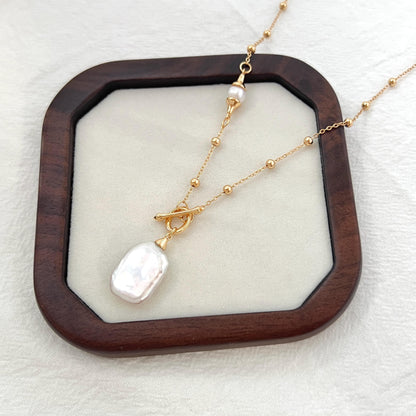 May include: A gold necklace with a large, irregular-shaped white pearl pendant. The necklace features a toggle clasp and small gold beads along the chain. A smaller pearl is also present near the top of the necklace. The jewelry is displayed on a light-colored surface within a dark wooden frame.